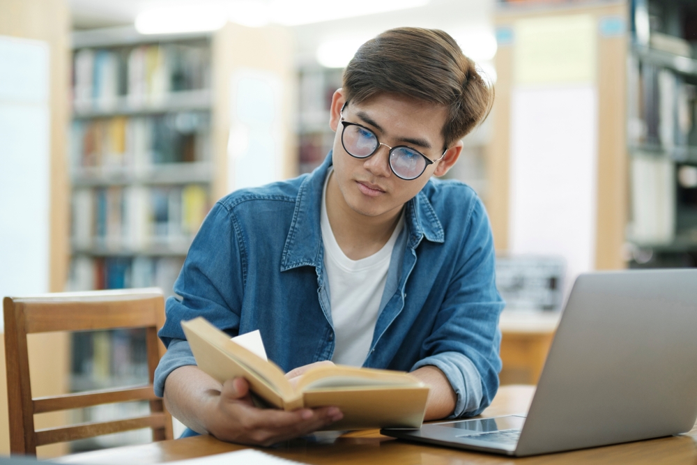 Young male college student wearing eyeglasses and in casual cloths studying and reading books using laptop at library for research or school project.