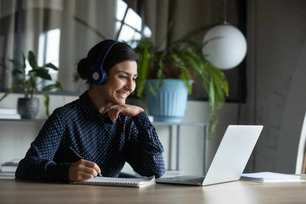 Happy young indian girl with wireless headphones looking at laptop screen, reading listening online courses, studying remotely from home with convenience and flexibility