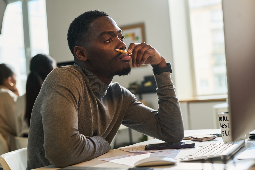 Young bored or tired male designer or broker with pencil by his mouth sitting in front of computer and looking at screen in office