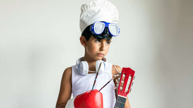 Kid wearing chef hat, swimming glasses headphones, paint brushes, boxing gloves and guitar represents child's passions and priorities